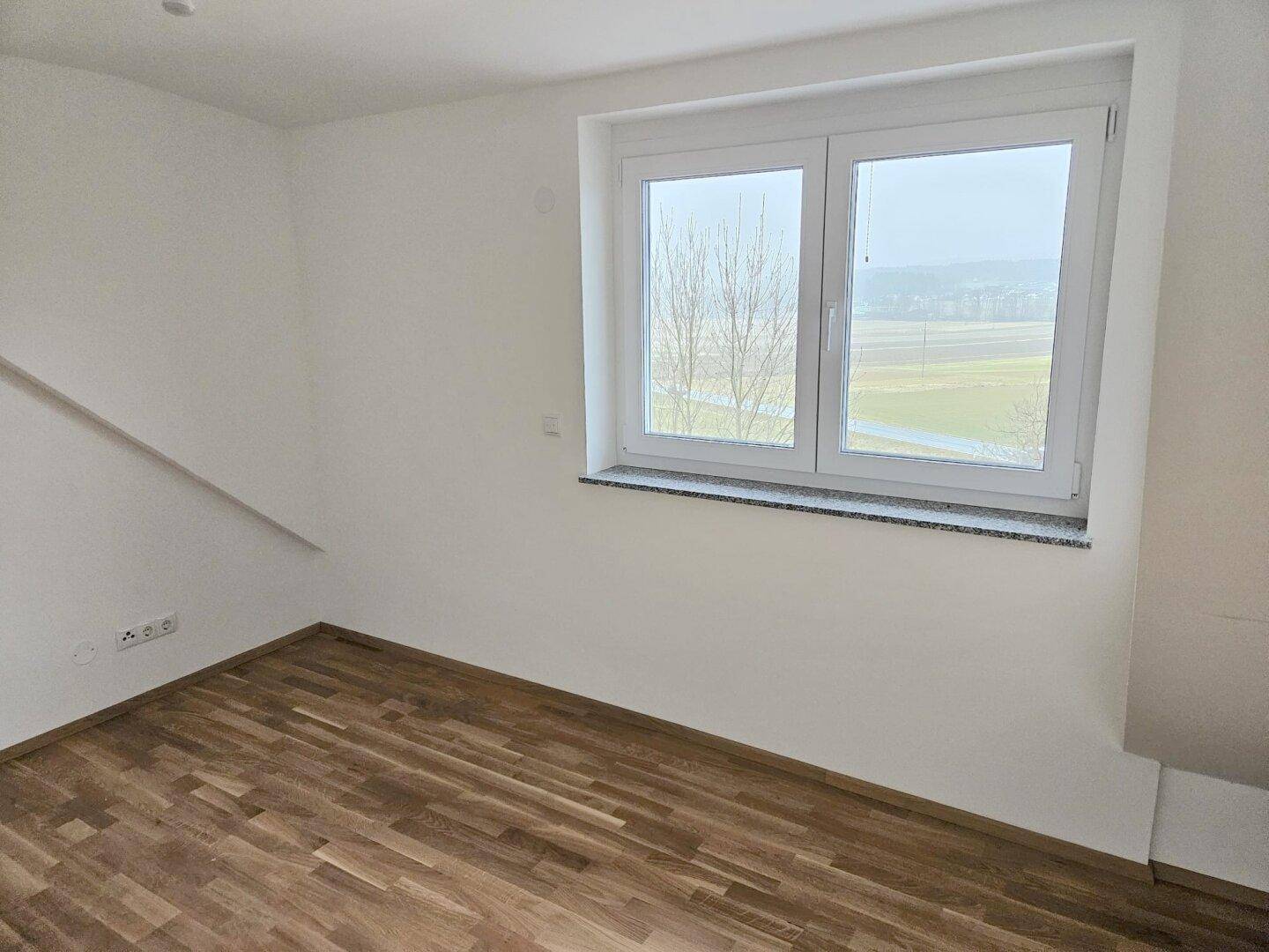Helles Zimmer mit Holzboden und einem Fenster, das einen weiten Blick über Felder bietet.