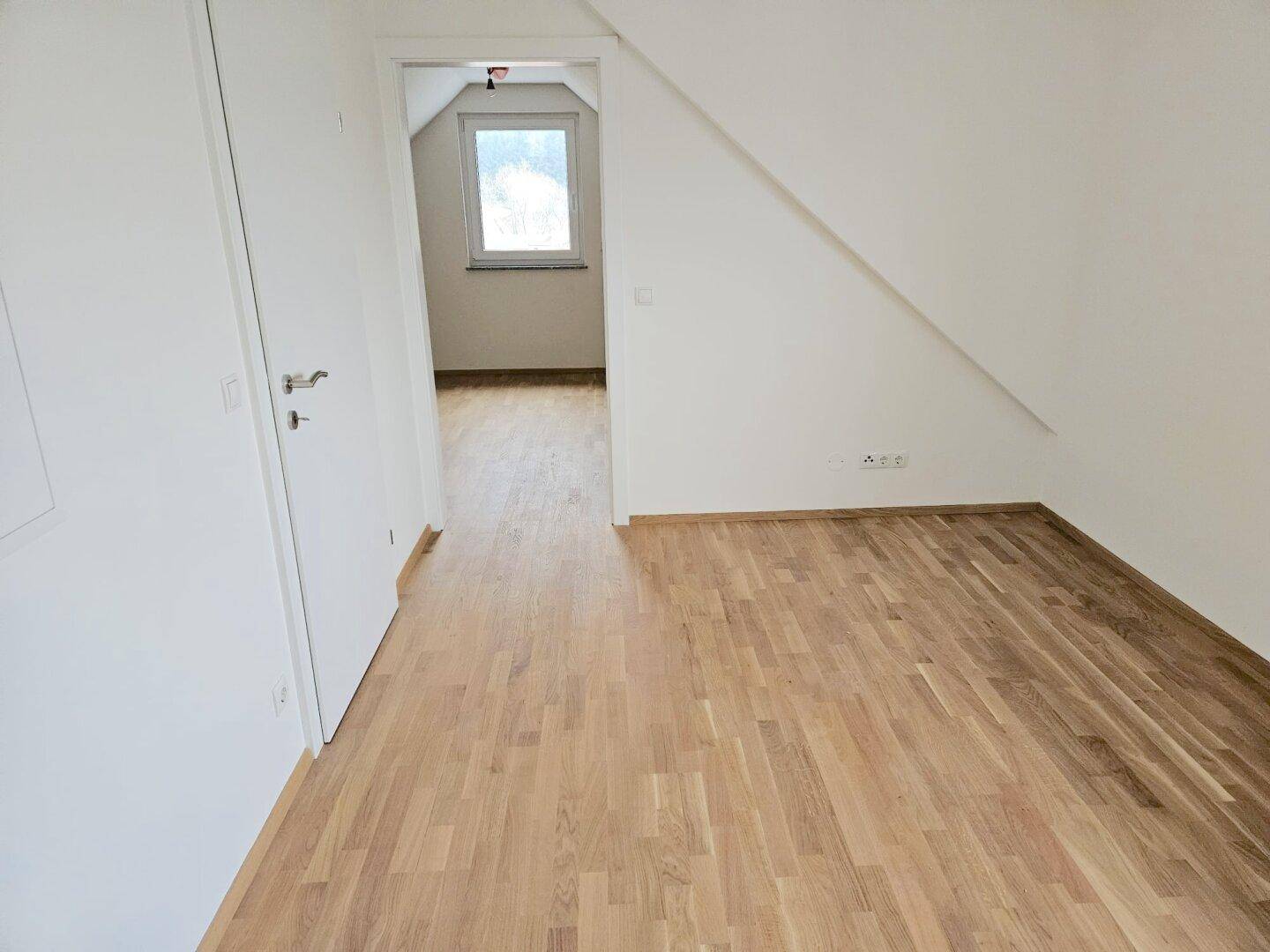 Heller Raum mit neuem Holzboden und Dachschrägen, der zu einem weiteren Zimmer führt.