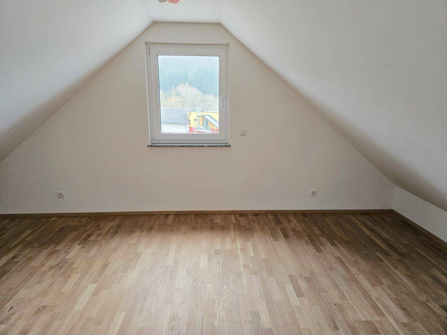 Geräumiges Zimmer im Dachgeschoss mit Holzboden, Dachschrägen und einem Fenster mit Blick ins Grüne.