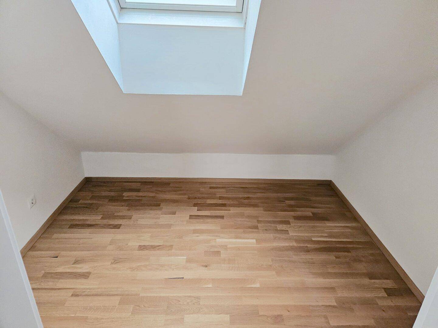 Leerer Raum im Dachgeschoss mit Holzboden, Dachschrägen und einem großen Dachfenster.