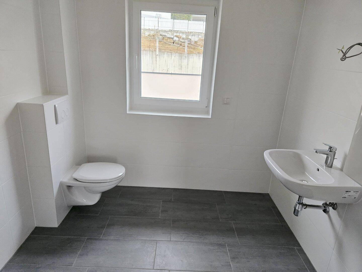 Modernes Badezimmer mit weißen Fliesen, dunklem Boden, wandhängendem WC und einem Fenster.