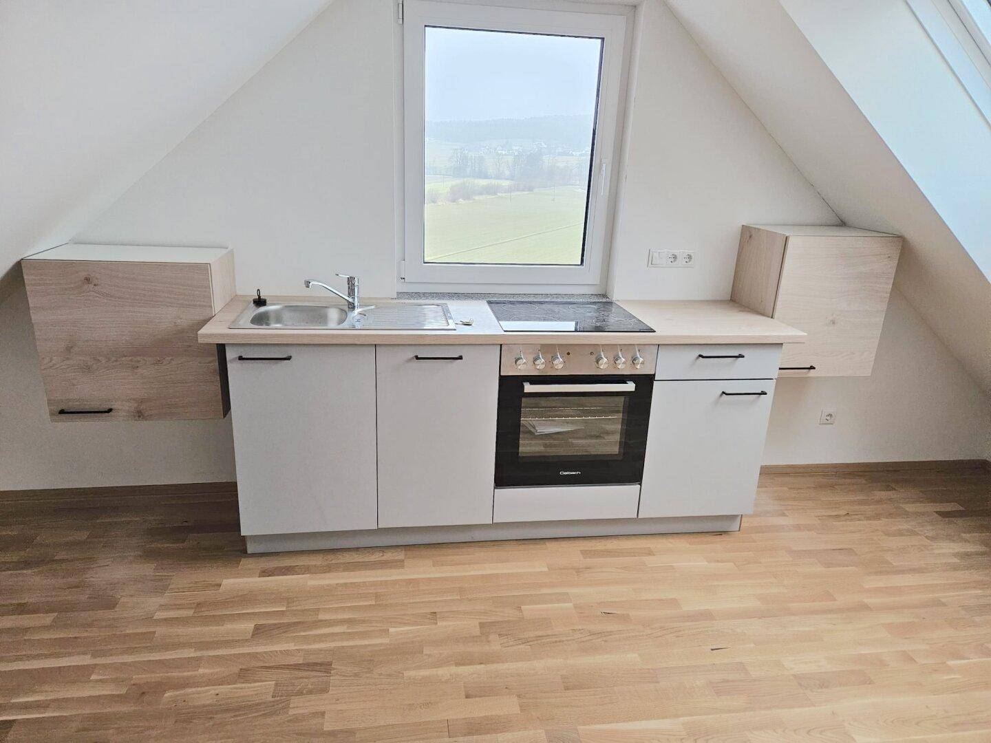 Kompakte Küchenzeile mit Kochfeld und Backofen in einem hellen Raum mit Dachschrägen und weitem Ausblick.
