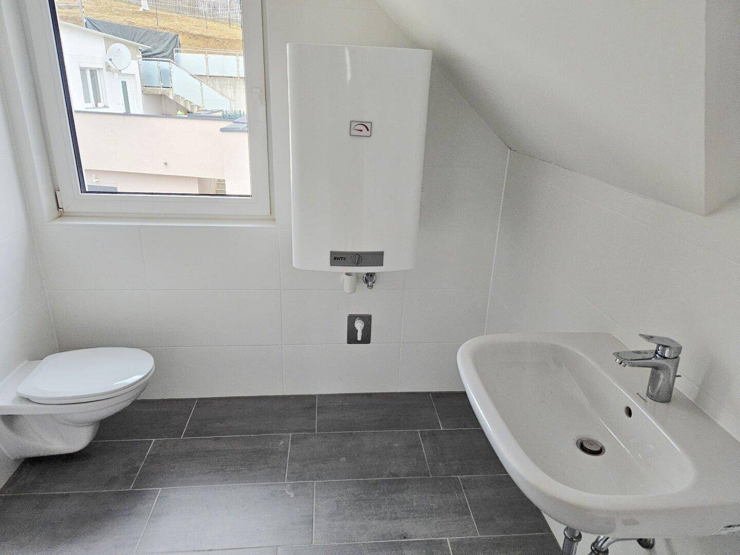 Helles Badezimmer mit Toilette, Waschbecken und Warmwasserboiler, ausgestattet mit weißen Wand- und grauen Bodenfliesen.