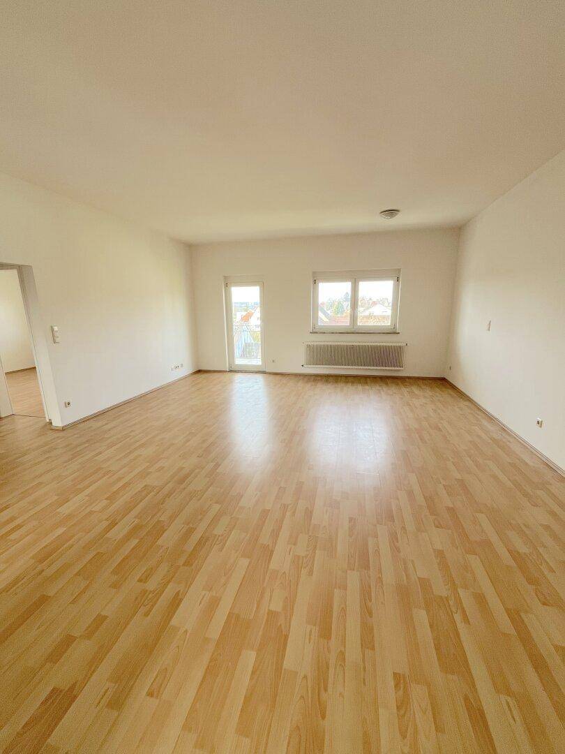 Helles, leeres Wohnzimmer mit hellem Laminatboden, weißen Wänden und Zugang zum Balkon.