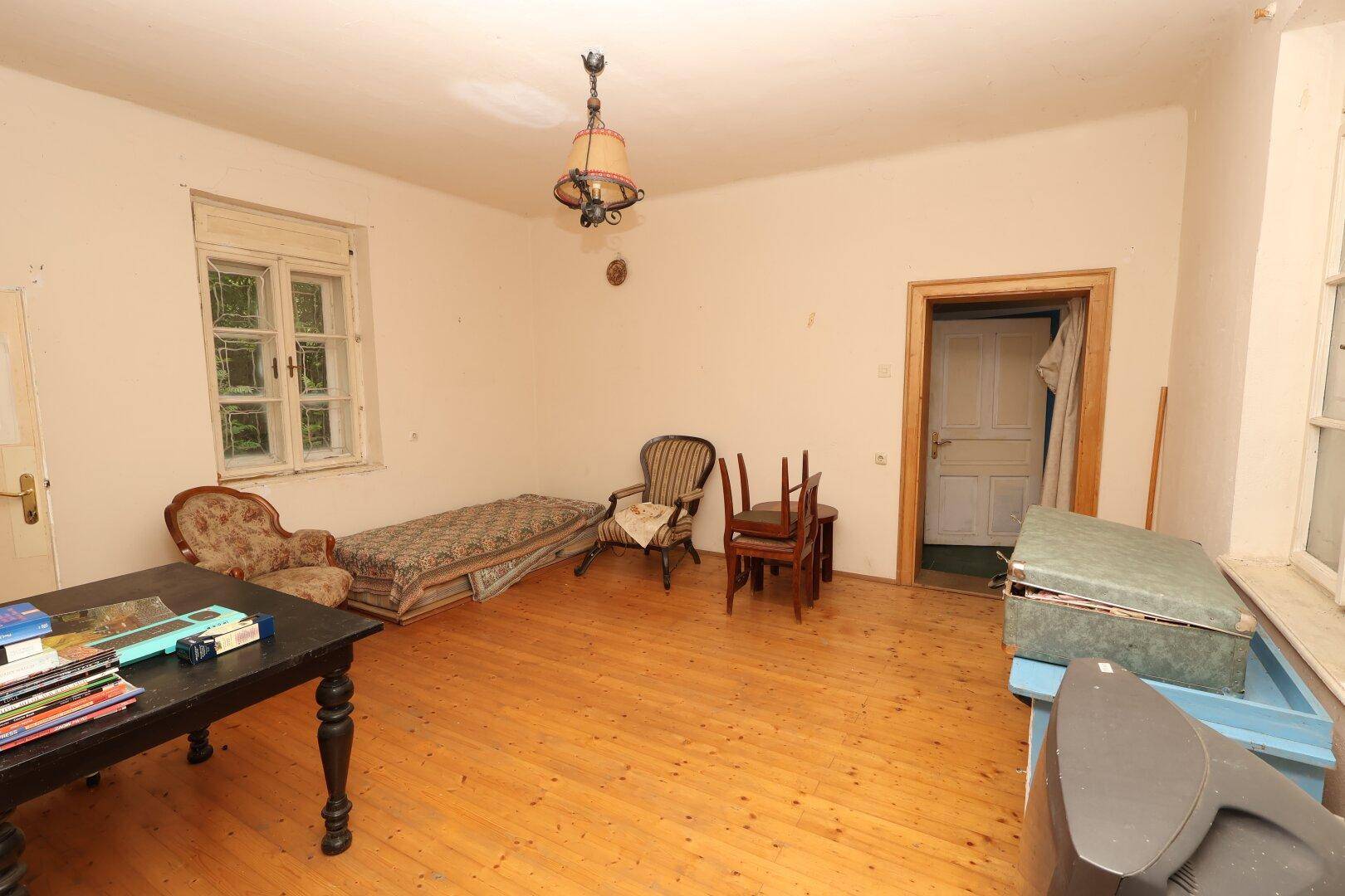 Geräumiges Wohnzimmer mit originalen Holzdielen, einem Fenster und einfacher, traditioneller Einrichtung, die auf Renovierungsbedarf hindeutet.