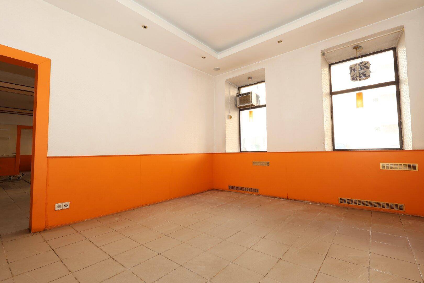 Geräumiger Raum mit Fliesenboden und orangefarbenem Anstrich, ausgestattet mit zwei Fenstern für Tageslicht.