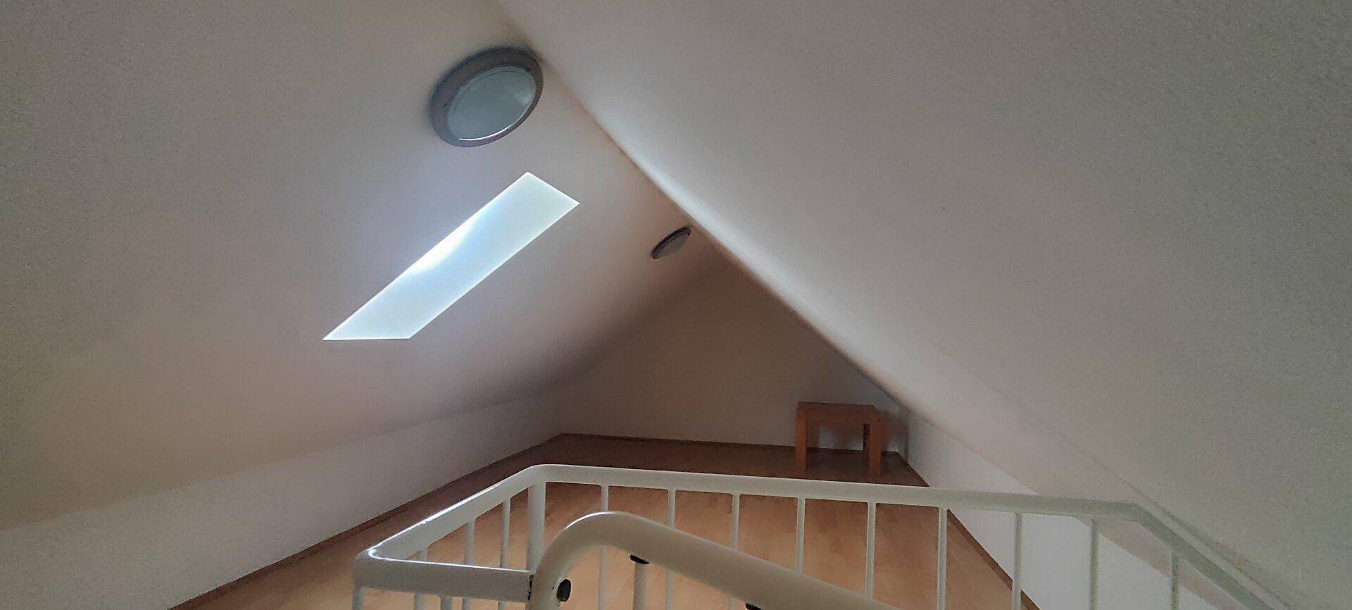 Dachgeschossbereich mit schrägen Wänden, einem Oberlicht und Blick auf eine Treppe.