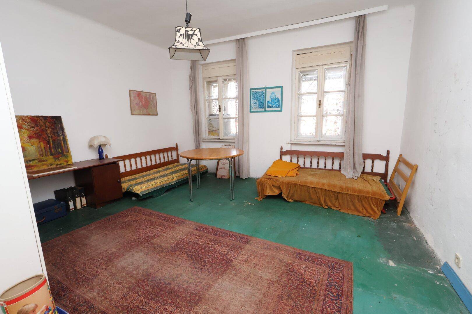 Geräumiges Zimmer mit grünem Boden, zwei Fenstern, Tagesbetten und einem großen gemusterten Teppich.