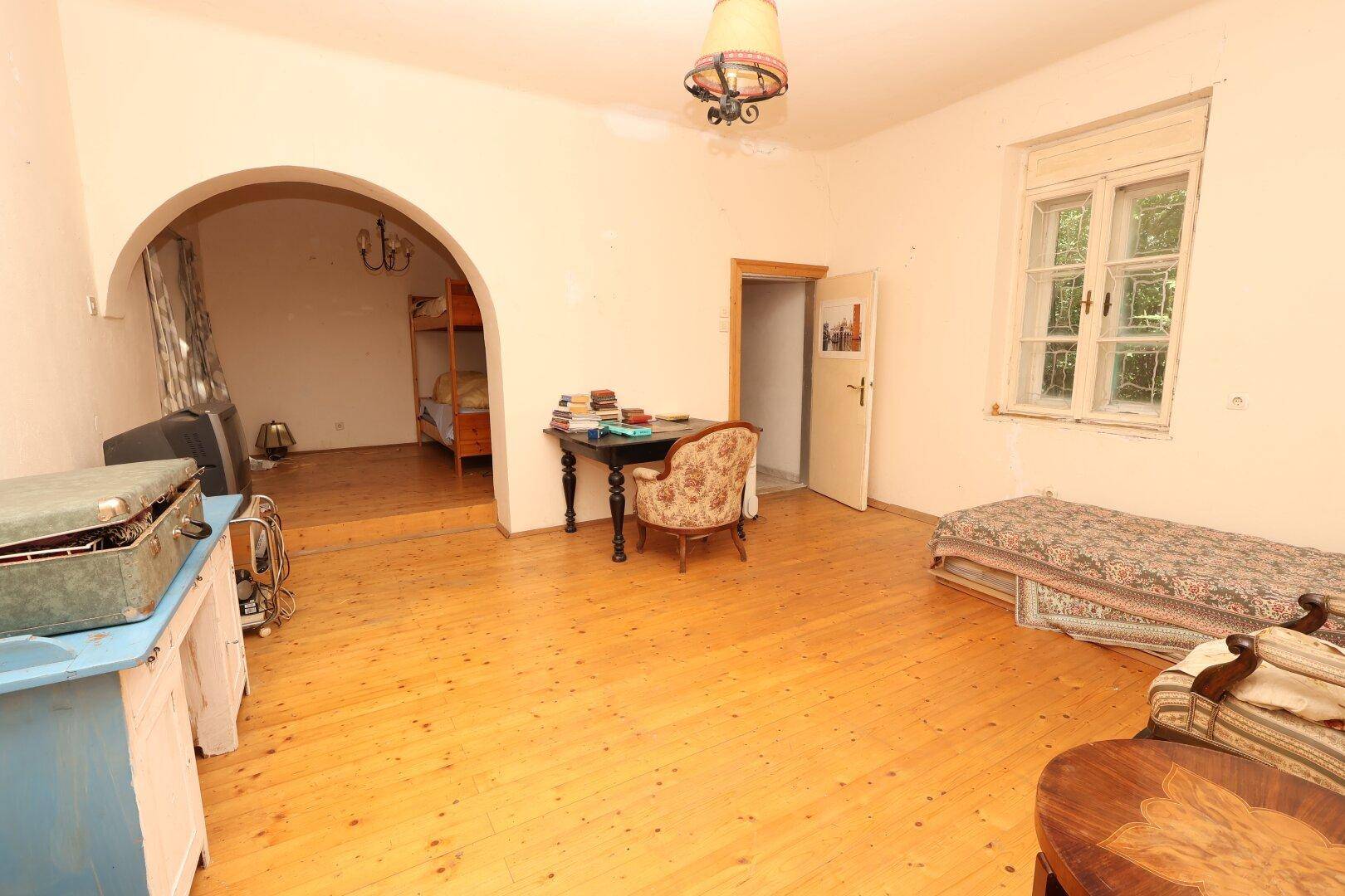 Helles Wohnzimmer mit Holzdielenboden, einem Bogen zu einem weiteren Raum und einem Fenster mit Blick ins Grüne.
