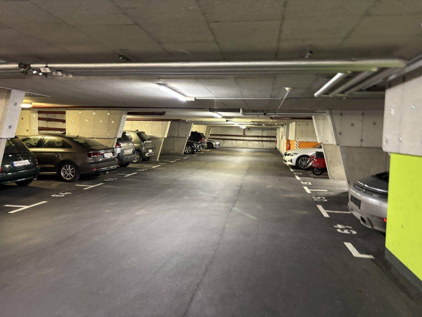 Lange Tiefgarage mit vielen Parkplätzen, Betonstützen und sichtbaren Rohren an der Decke.