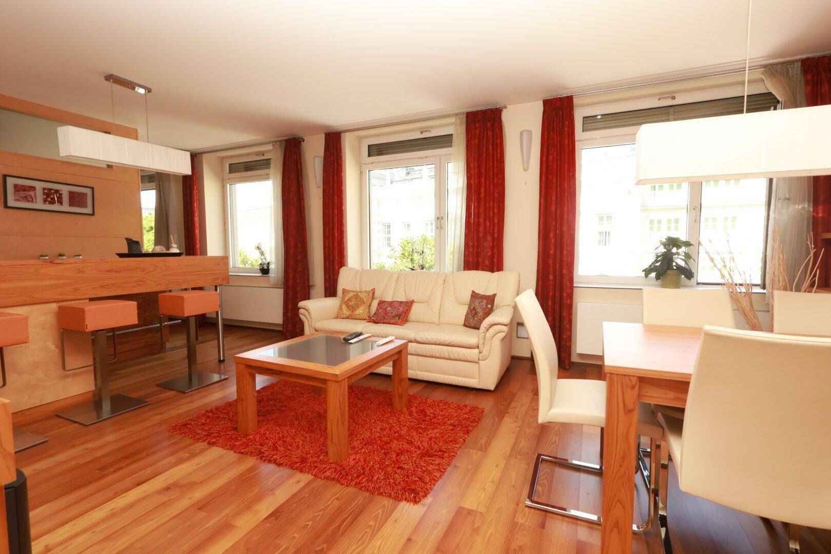 Heller offener Wohn- und Essbereich mit Holzboden, beigem Sofa und rotem Teppich sowie einer Bar.