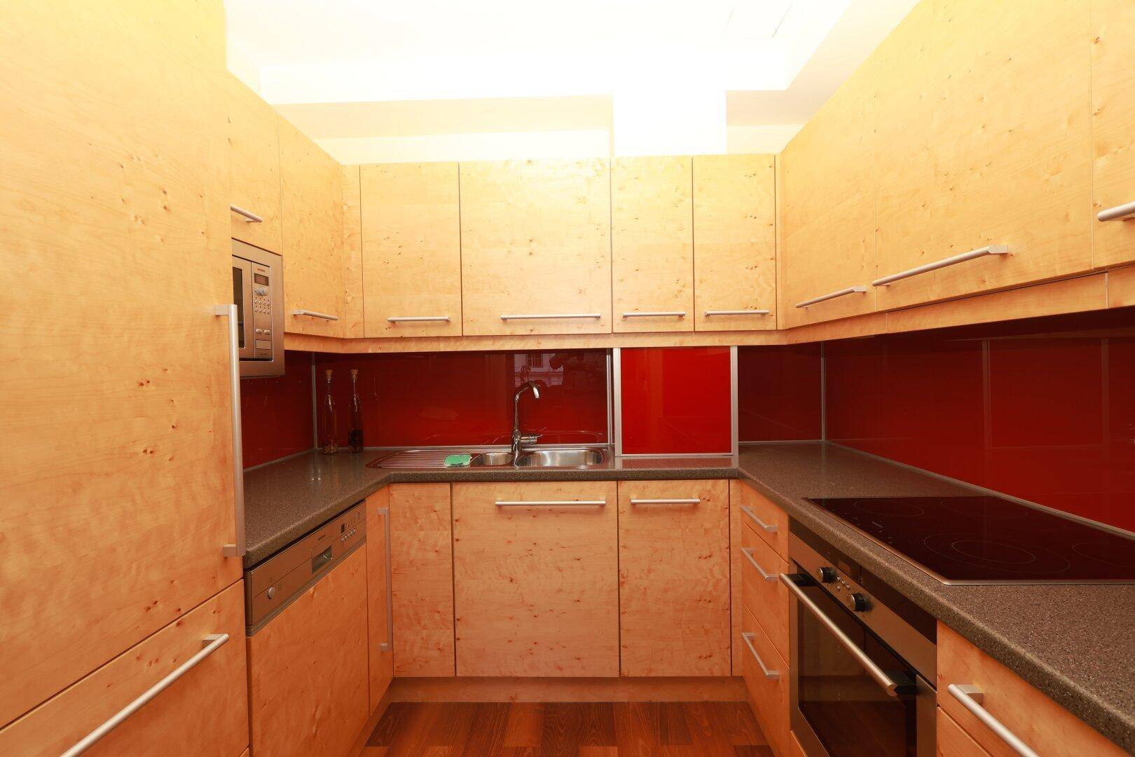 Moderne Einbauküche mit hellen Holzfronten, dunkler Arbeitsplatte und roter Rückwand.