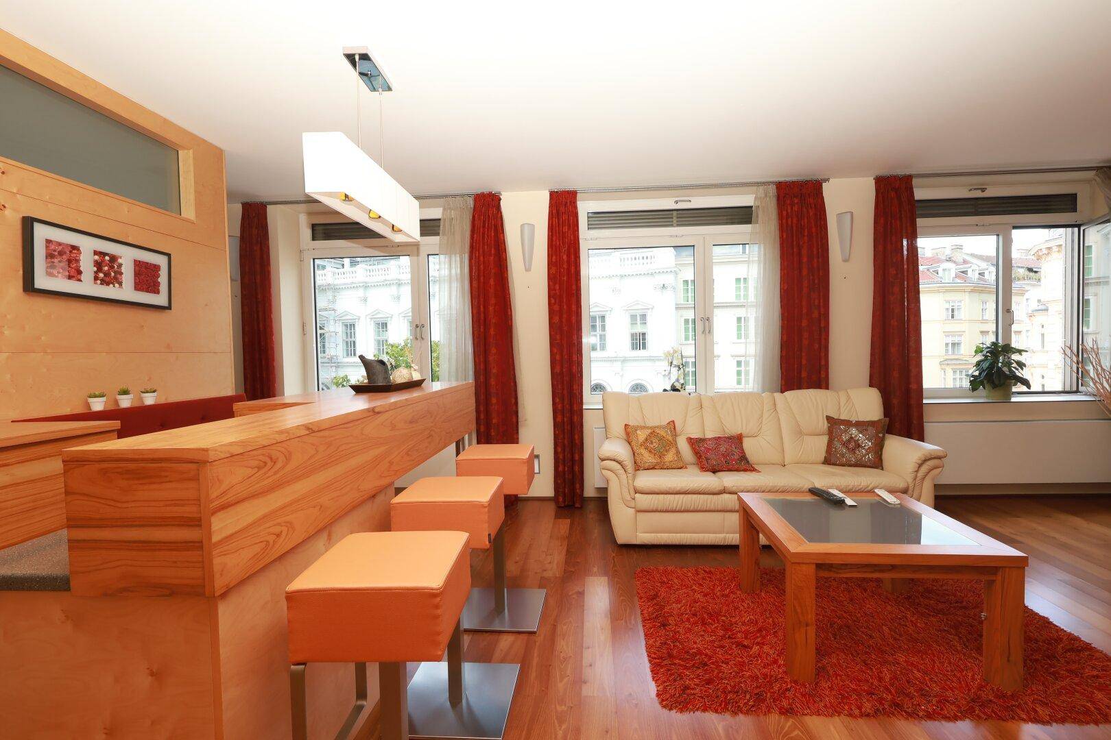 Heller Wohn- und Essbereich mit Holzboden, beigem Sofa und rotem Teppich sowie einer Bar.