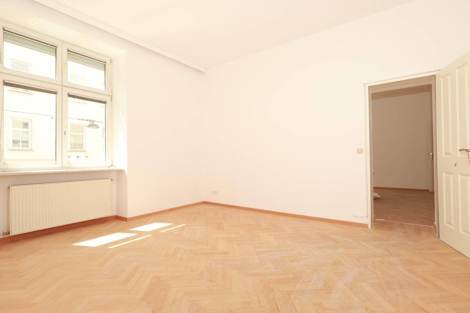 Geräumiges Zimmer mit hellem Fischgrätparkett, weißen Wänden und einem Fenster mit Blick nach draußen.