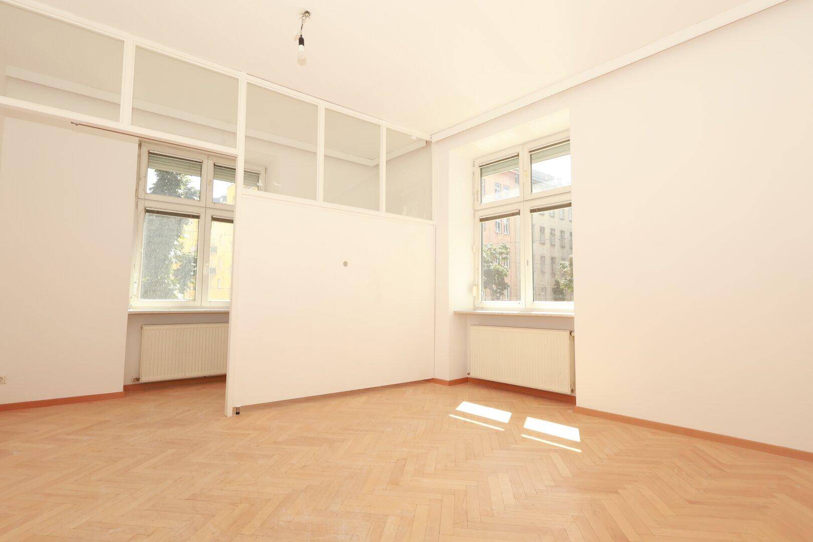 Helles Zimmer mit Fischgrätparkett, weißen Wänden und einer modernen Glasabtrennung, die den Raum teilt.