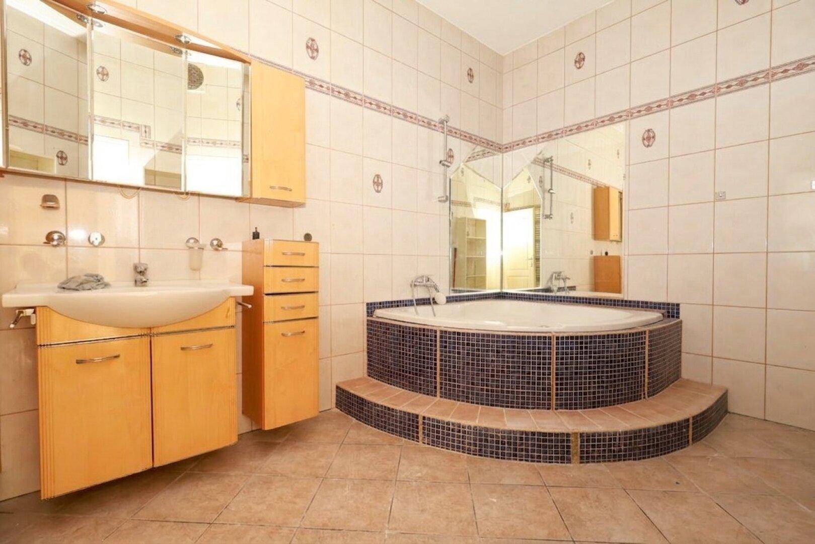 Badezimmer mit Eckbadewanne, Einzelwaschbecken und beigefarbenen Fliesen, akzentuiert durch blaue Mosaikfliesen.