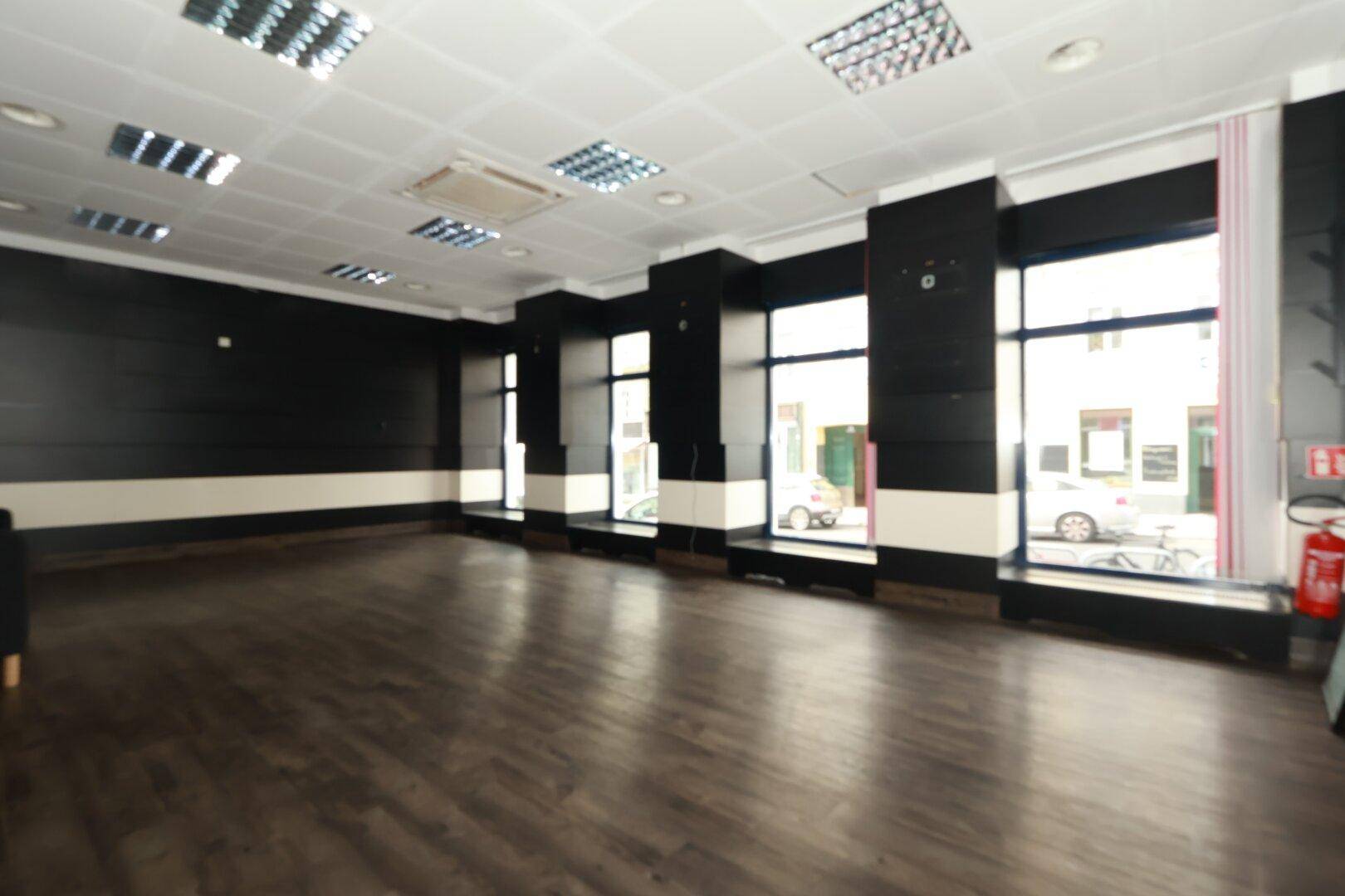 Großer, leerer Geschäftsraum mit dunklem Holzboden, weißen Deckenplatten und großen Schaufenstern zur Straße.
