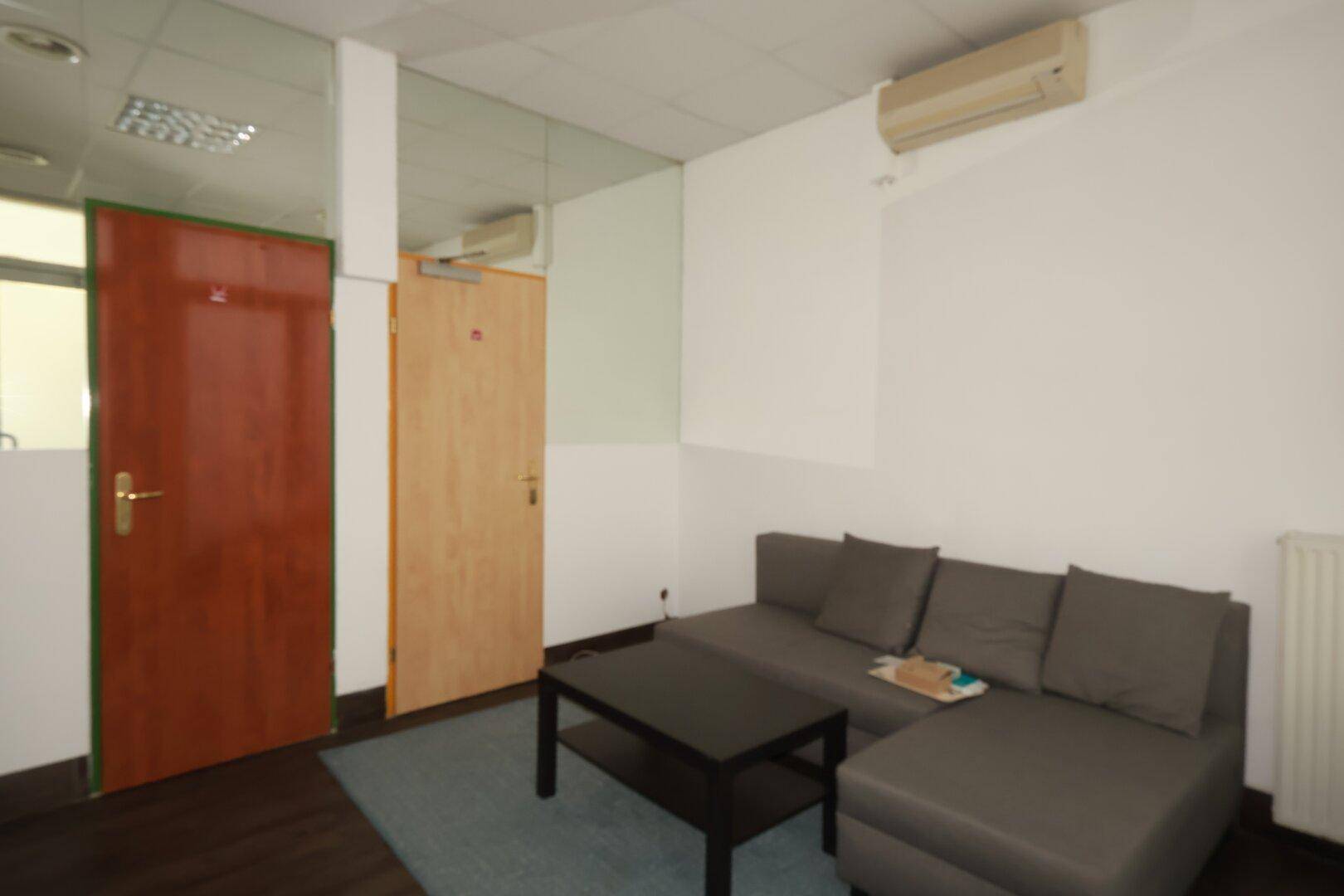 Kleiner Bürobereich mit Sofa, Couchtisch, zwei Türen und Klimaanlage an der Wand.