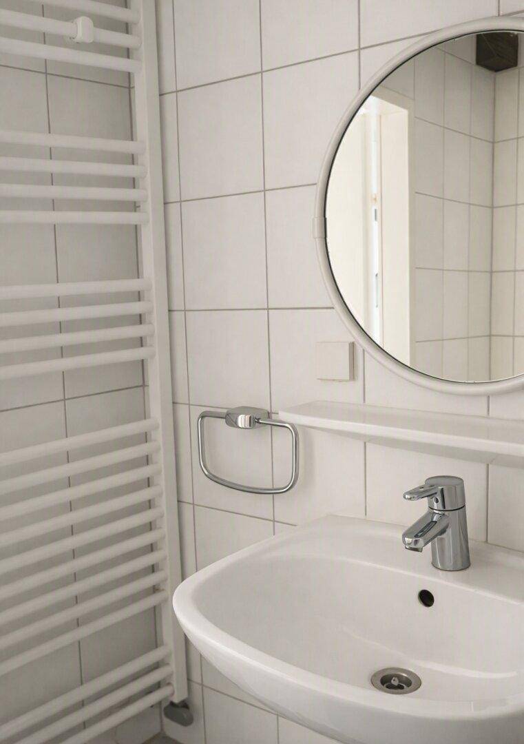Modernes Badezimmer mit weißem Waschbecken, rundem Spiegel und beheiztem Handtuchhalter.