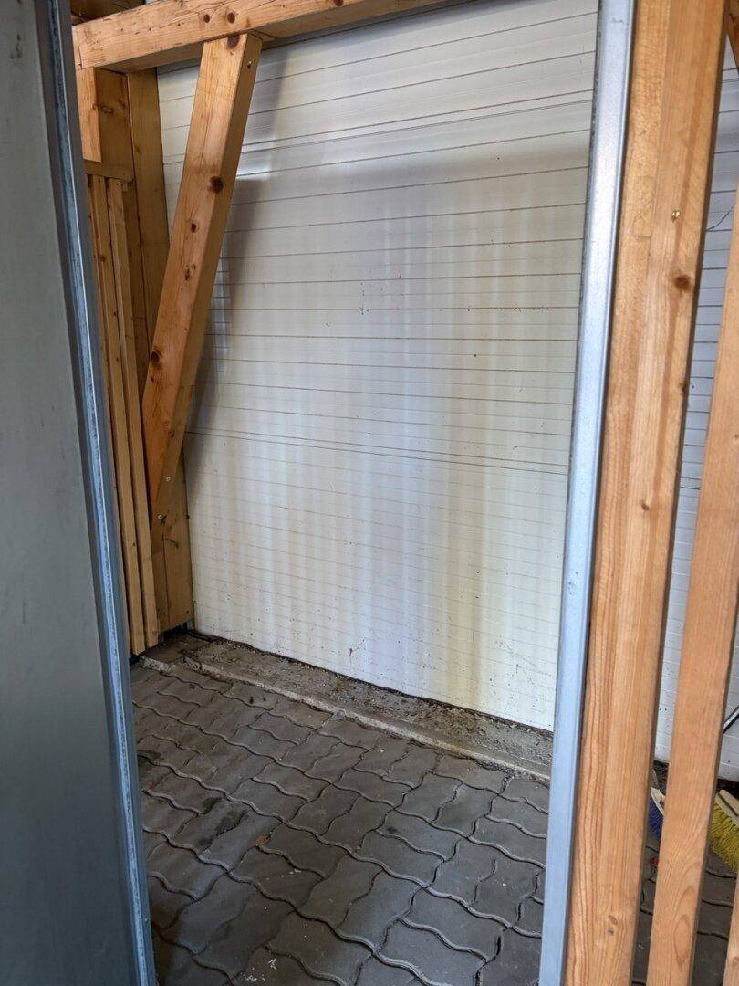 Einfacher Abstellraum mit Holzkonstruktion und gepflastertem Boden im Carportbereich.