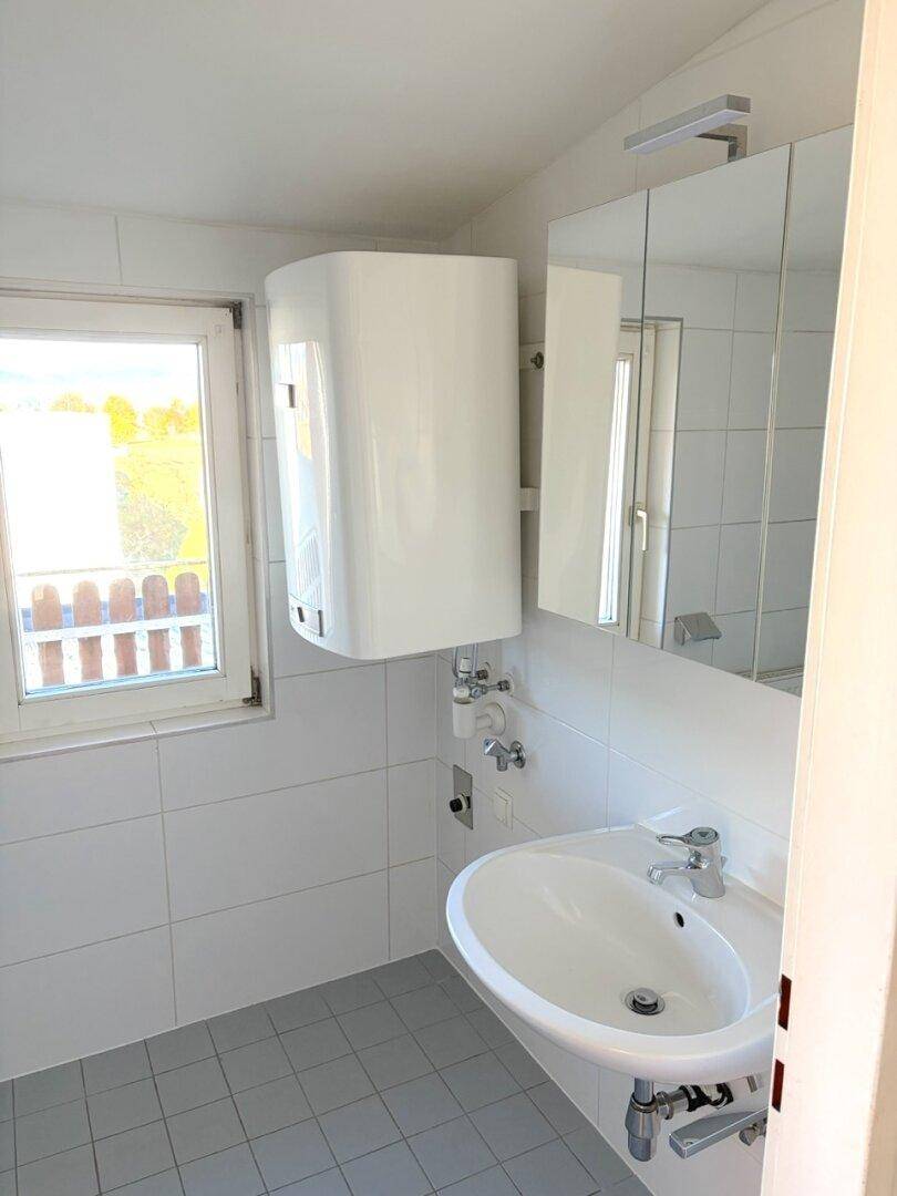 Helles Badezimmer mit Waschbecken, Spiegelschrank, Fenster und Warmwasserboiler.
