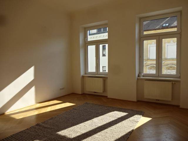 Heller Raum mit Fischgrätparkett, weißen Wänden, zwei Fenstern und einem Teppich, durchflutet von Sonnenlicht.
