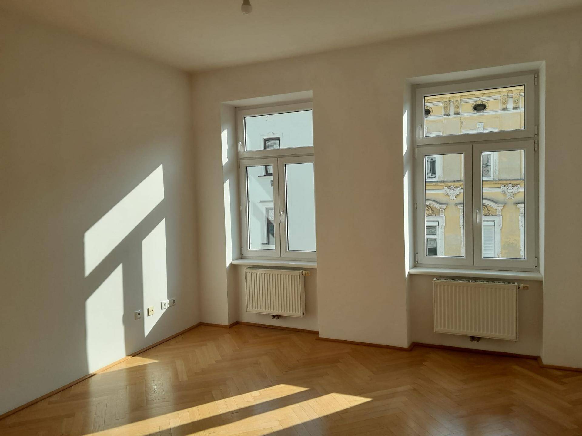 Heller Raum mit Fischgrätparkett, weißen Wänden und zwei großen Fenstern, durch die Sonnenlicht fällt.