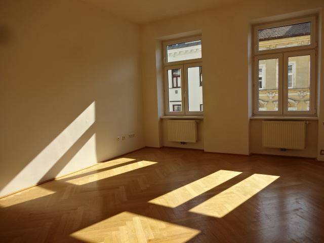 Heller Raum mit Fischgrätparkett, weißen Wänden und zwei großen Fenstern, durch die Sonnenlicht fällt.