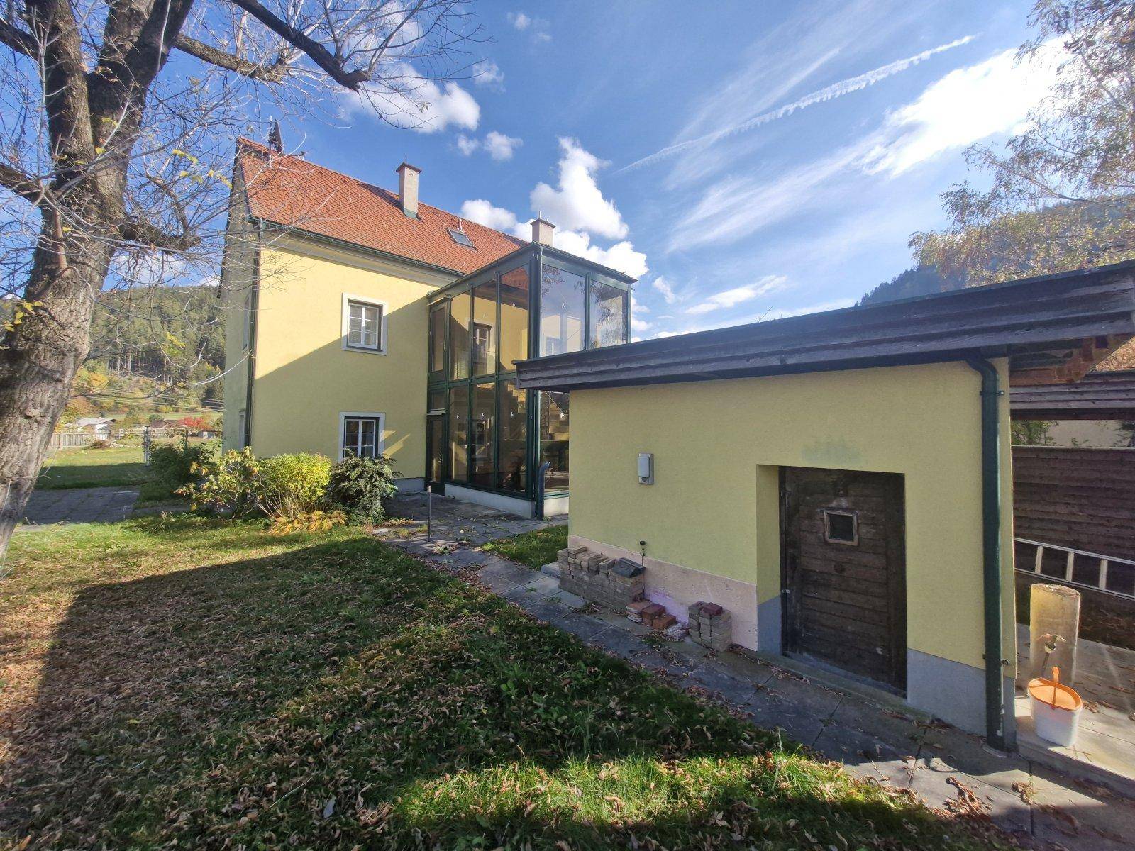Seitenansicht des Hauses mit Rasenfläche, kleinem Nebengebäude und dem verglasten Anbau.