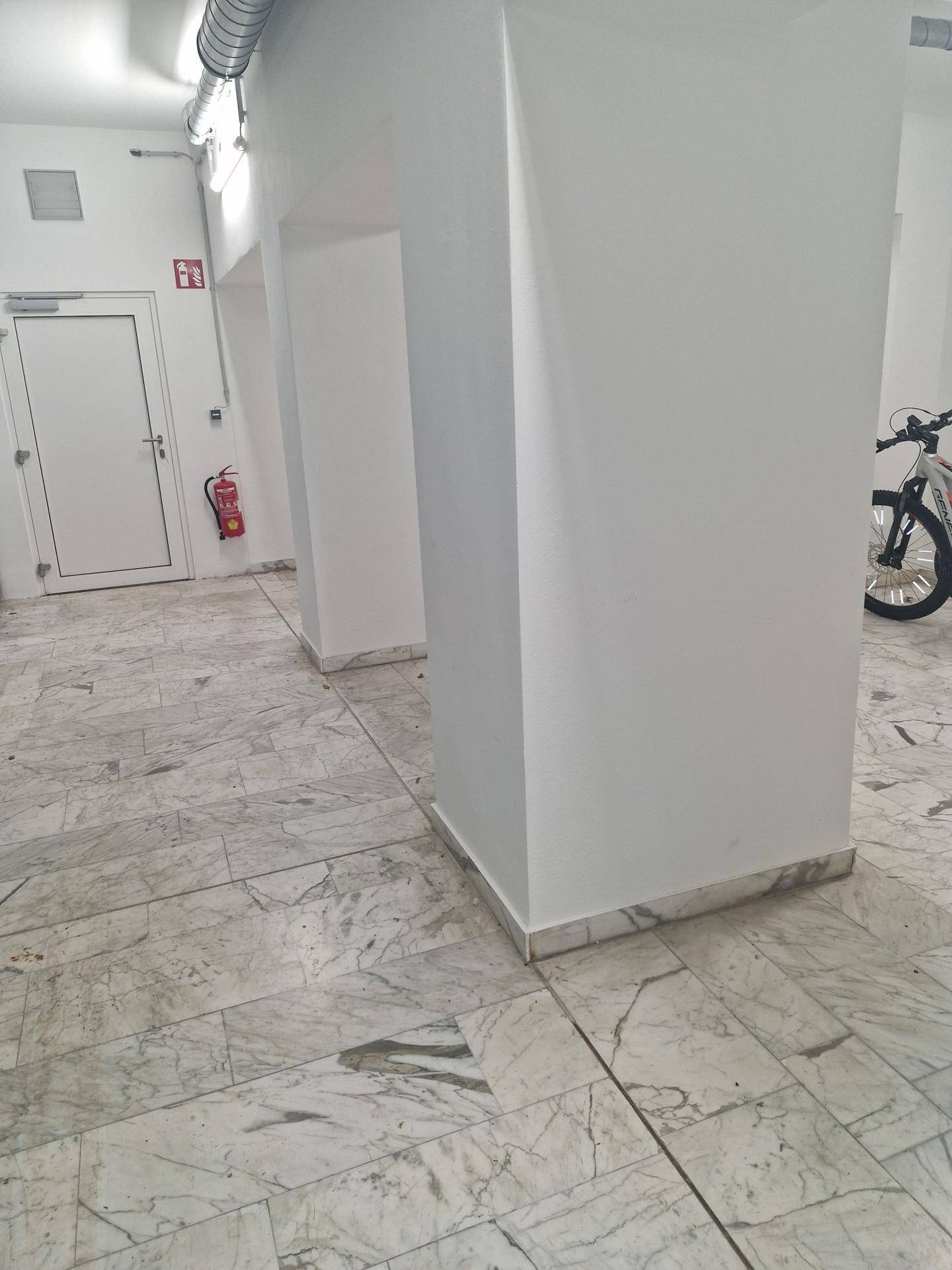 Langer Flur mit Marmorboden, weißen Wänden und sichtbaren Lüftungsrohren an der Decke.