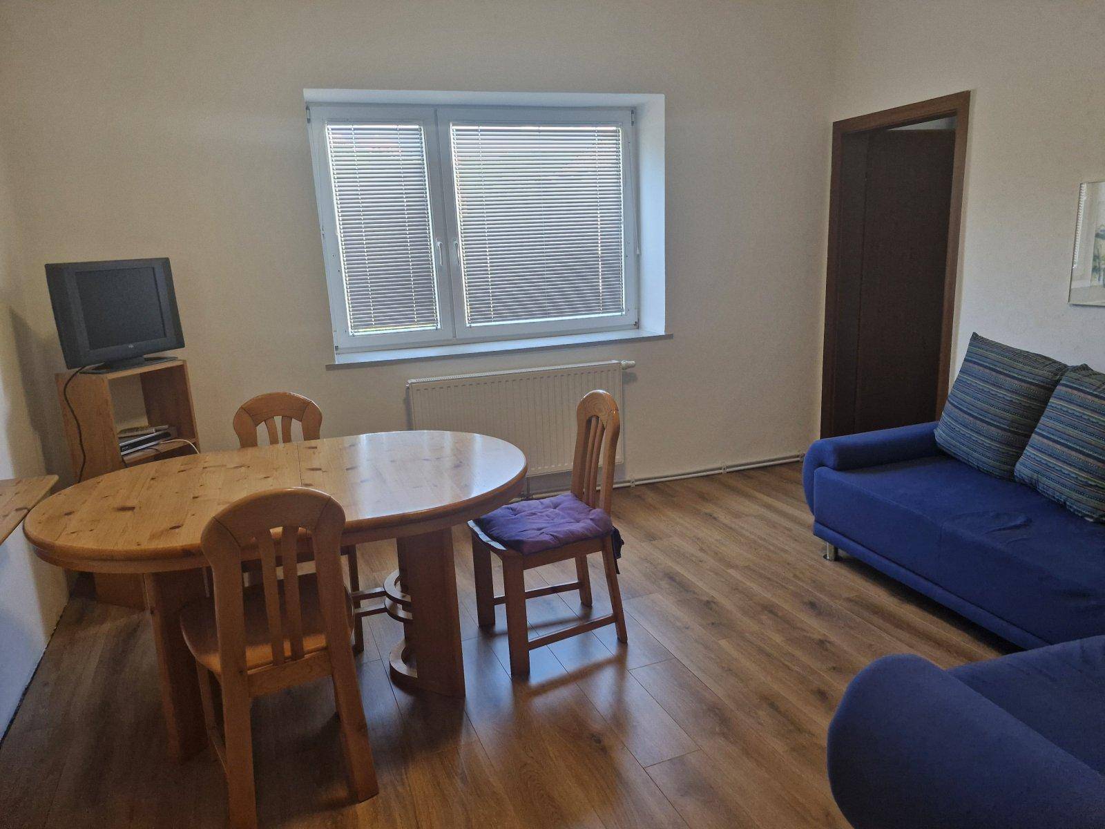 Gemütlicher Wohn-Essbereich mit Holztisch, blauer Couch und Fenster mit Jalousien.