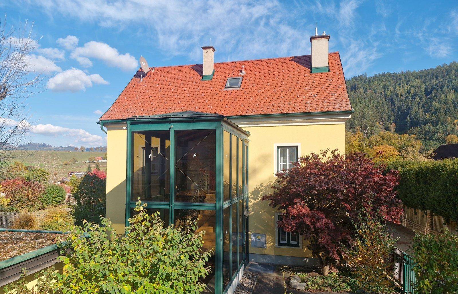 Gelbes Haus mit verglastem Wintergarten und rotem Ziegeldach vor bewaldeter Bergkulisse.