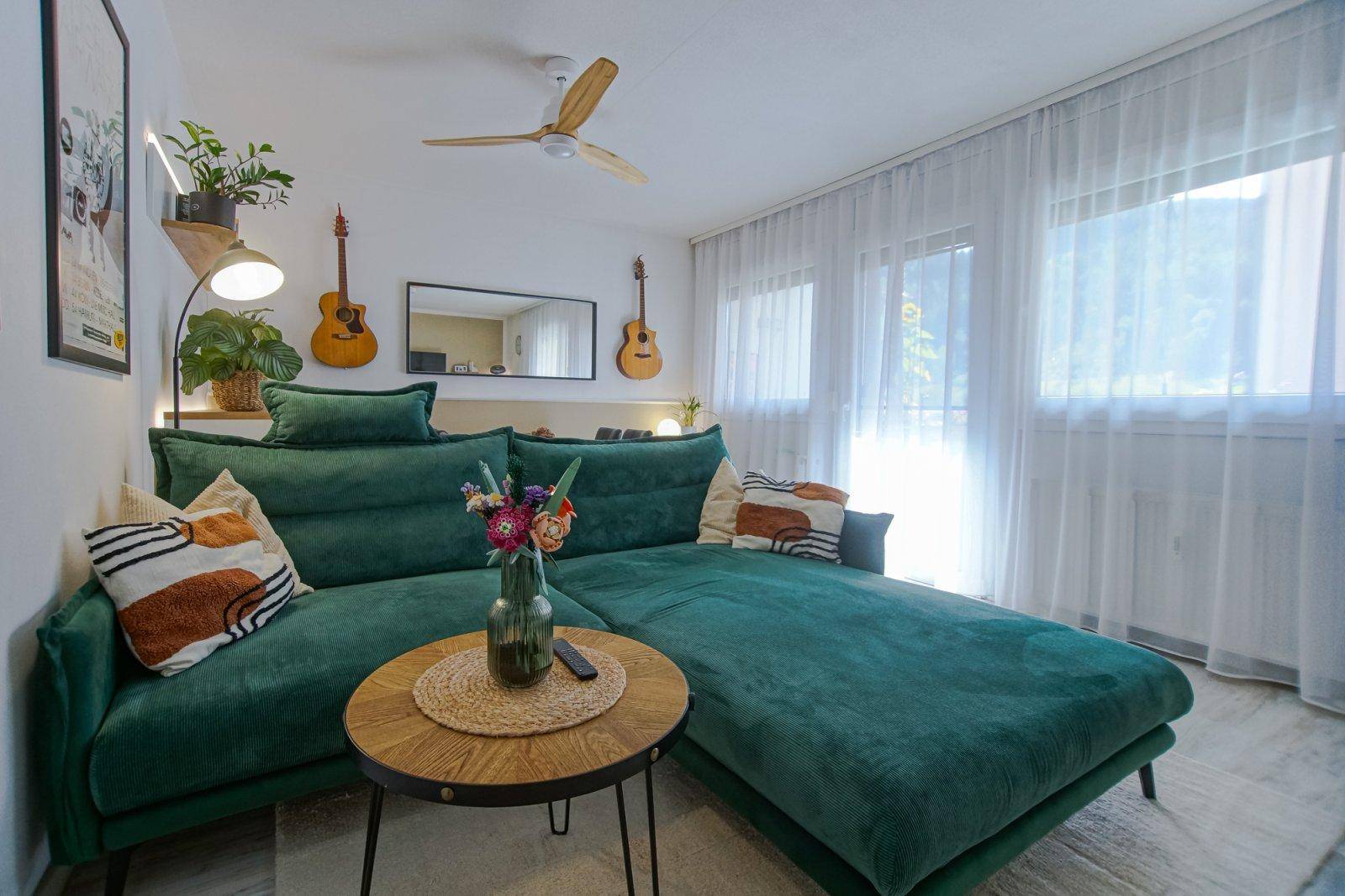 Gemütliches Wohnzimmer mit grüner Eckcouch, Deckenventilator und hellen Fenstern mit Bergblick.
