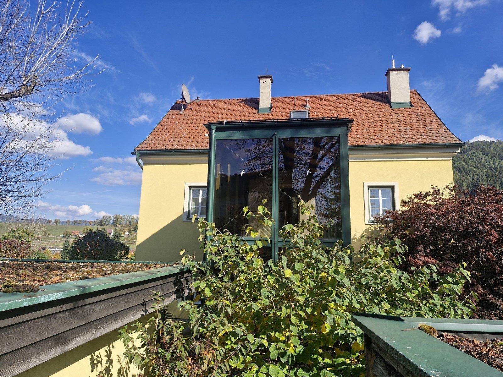 Blick über ein Gründach auf den verglasten Anbau des Hauses unter blauem Himmel.