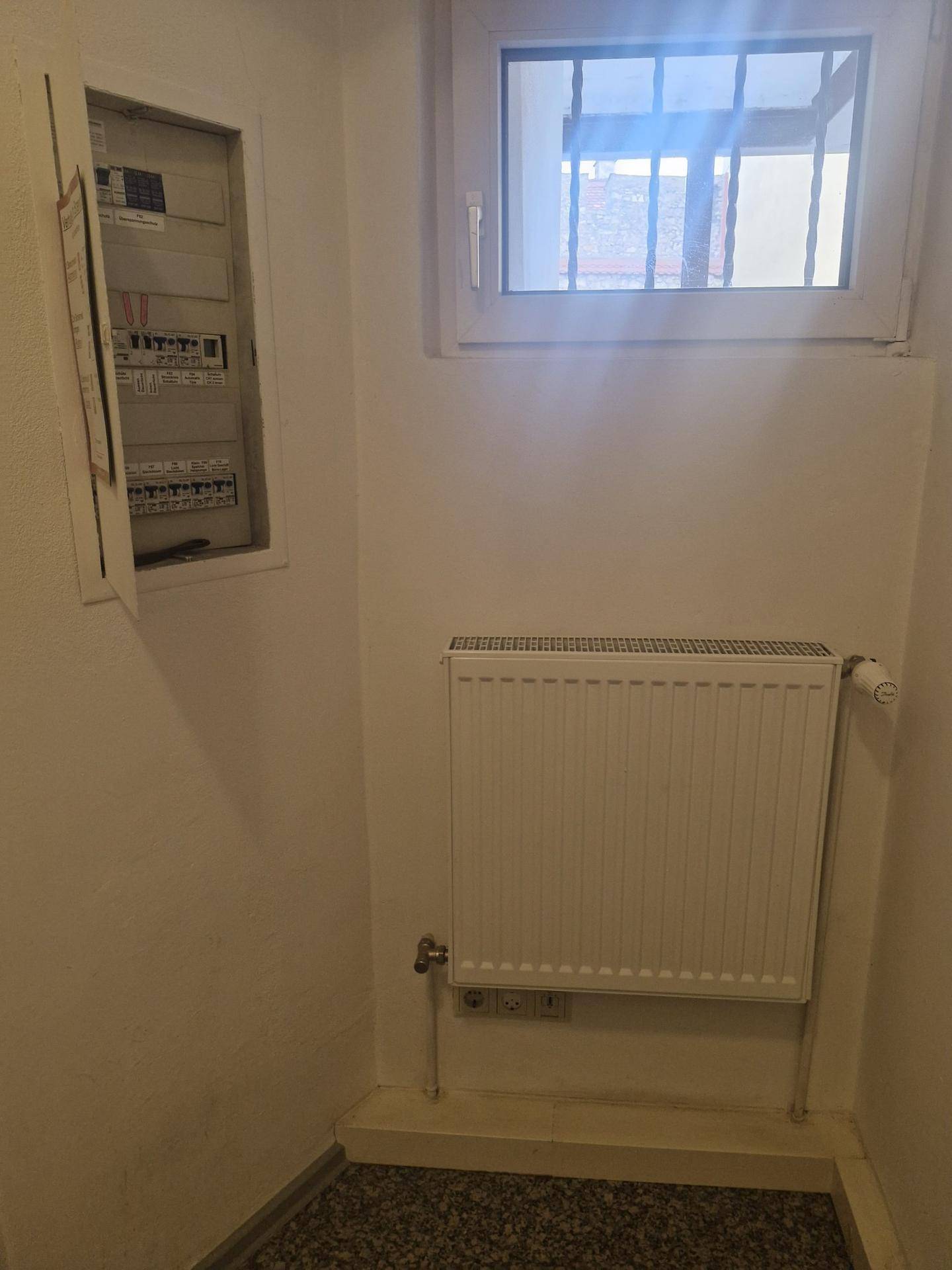 Kleiner Bereich mit einem Fenster, Heizkörper und einem geöffneten Sicherungskasten an der Wand.