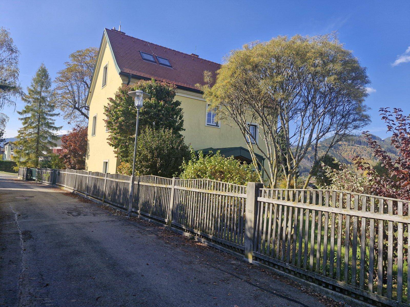 Seitenansicht des gelben Hauses von der Straße mit Holzzaun und Bäumen.