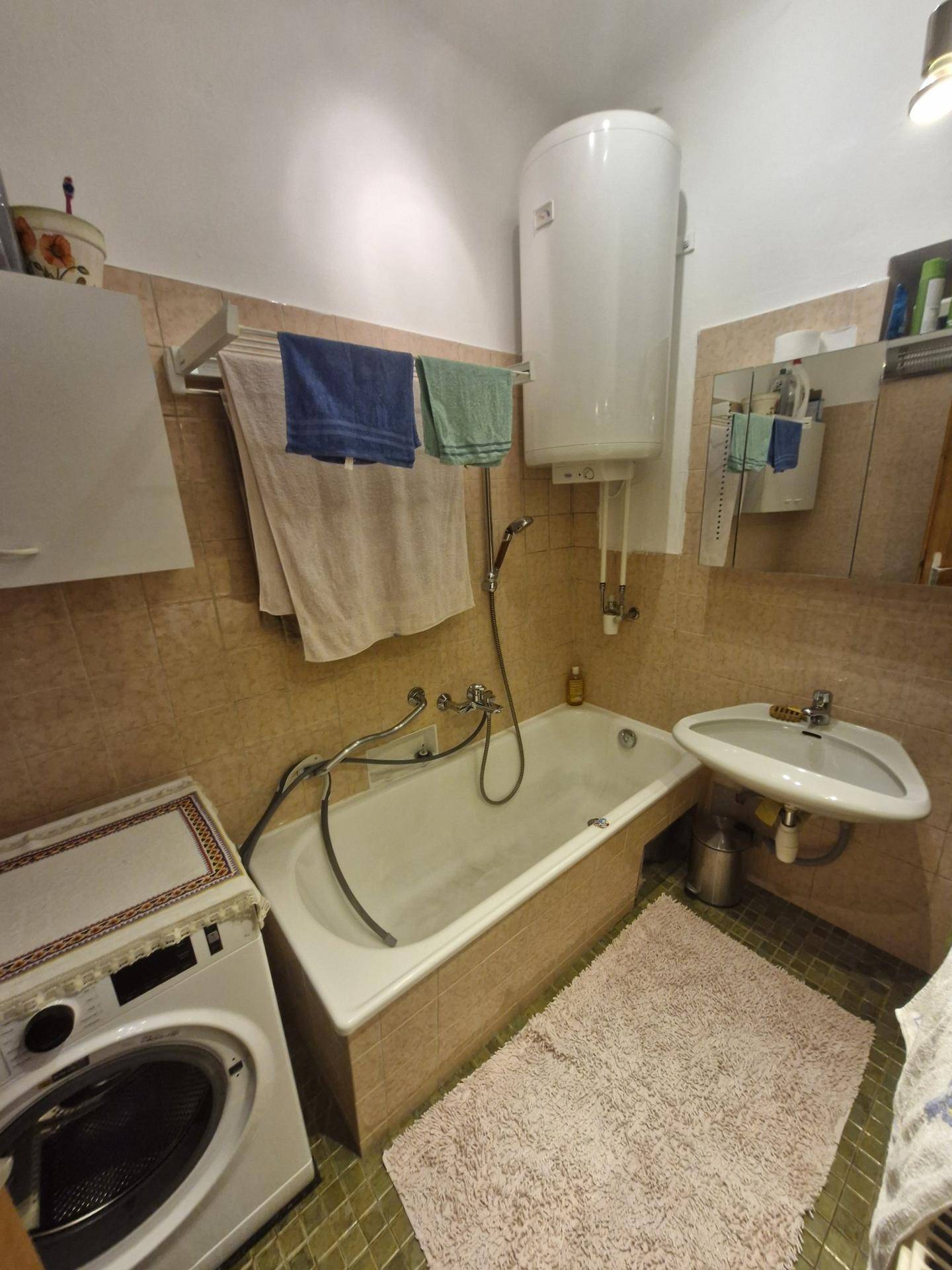 Badezimmer mit Badewanne, Waschmaschine und Boiler, gefliest in beigen Tönen.