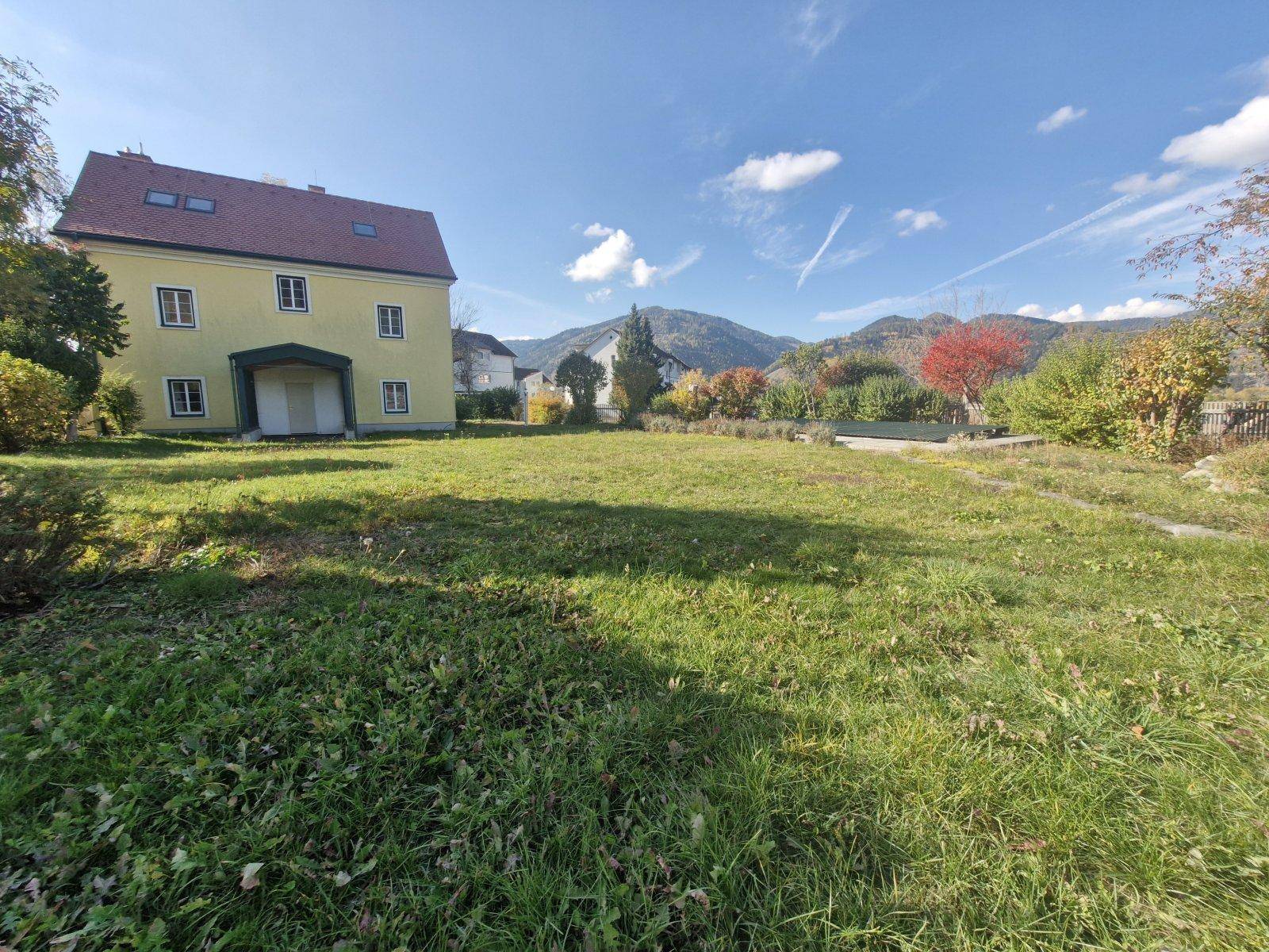 Weitläufige Rasenfläche vor dem gelben Haus mit Blick auf die umliegende Landschaft und Berge.