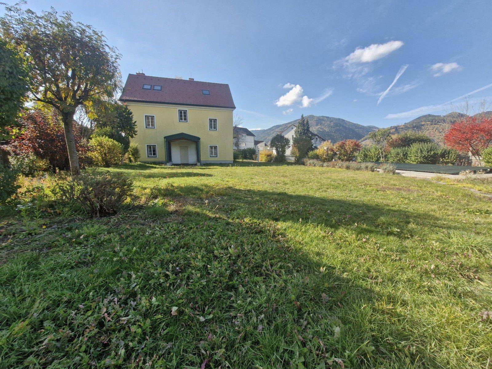 Weitläufige Rasenfläche vor dem gelben Haus mit Blick auf die umliegende Landschaft und Berge.