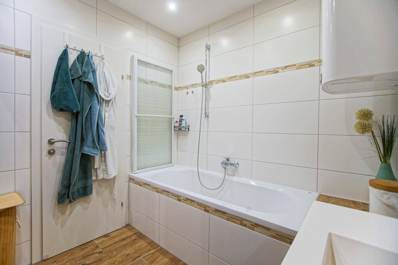 Badezimmer mit Badewanne, Duschfunktion und weißen Fliesen, ausgestattet mit einem Warmwasserboiler.