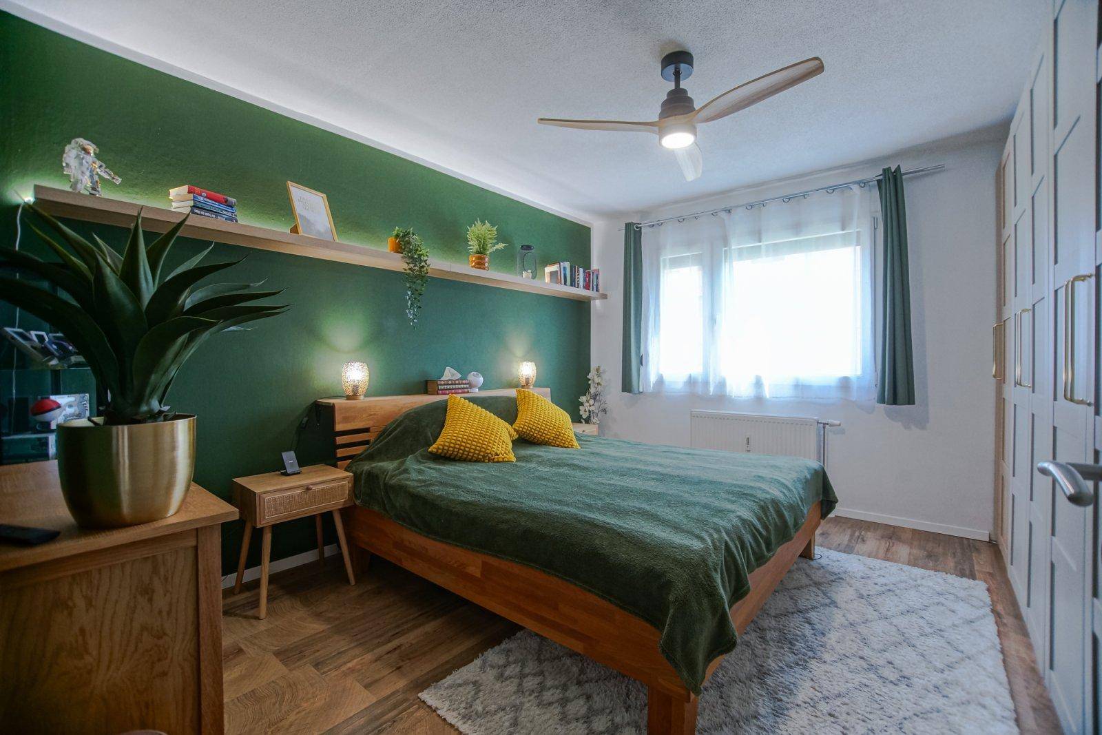 Gemütliches Schlafzimmer mit Holzbett, grüner Akzentwand, Deckenventilator und Fenster.