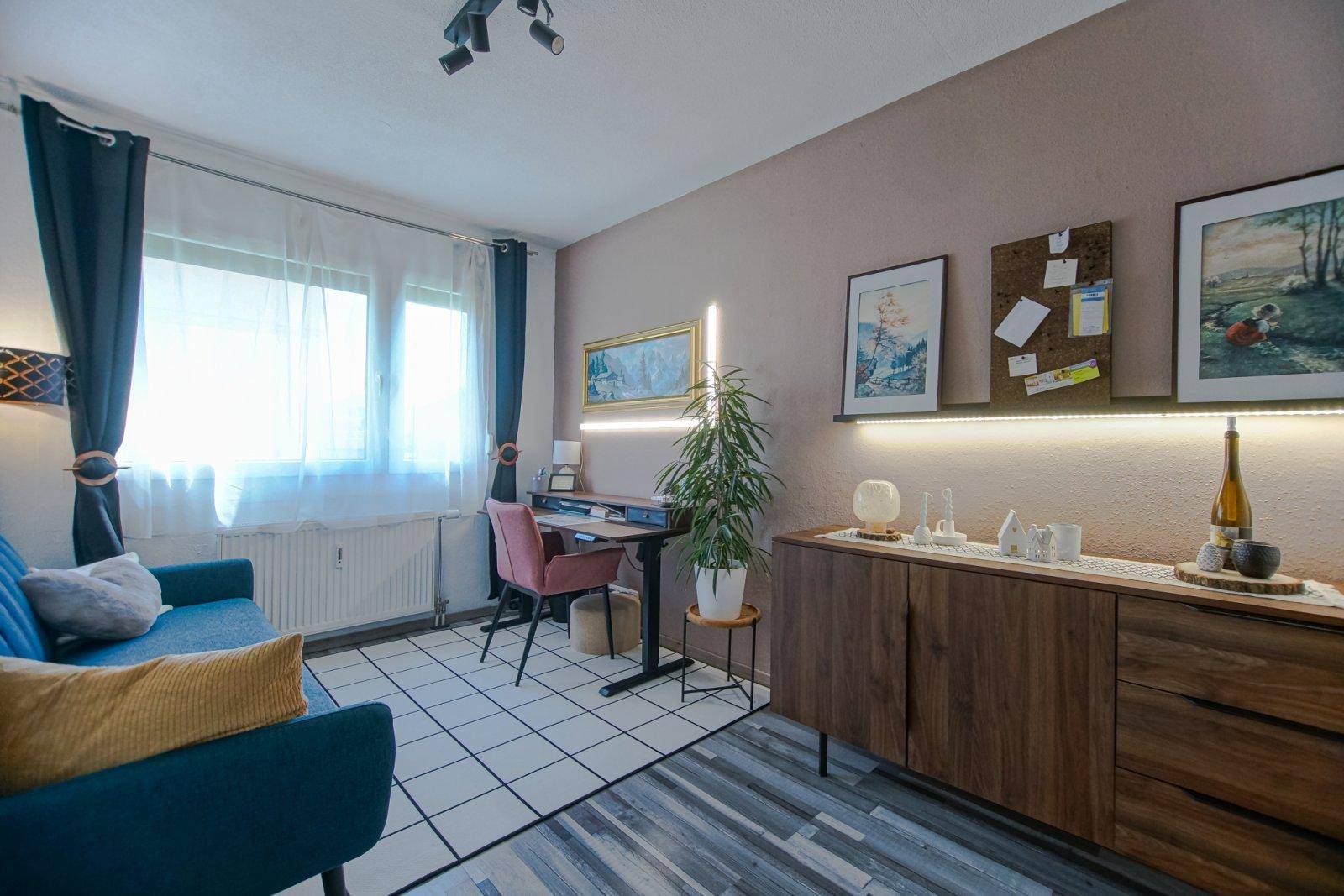 Helles Wohnzimmer mit Sofa, Schreibtisch und Sideboard, teilweise gefliestem und laminiertem Boden.