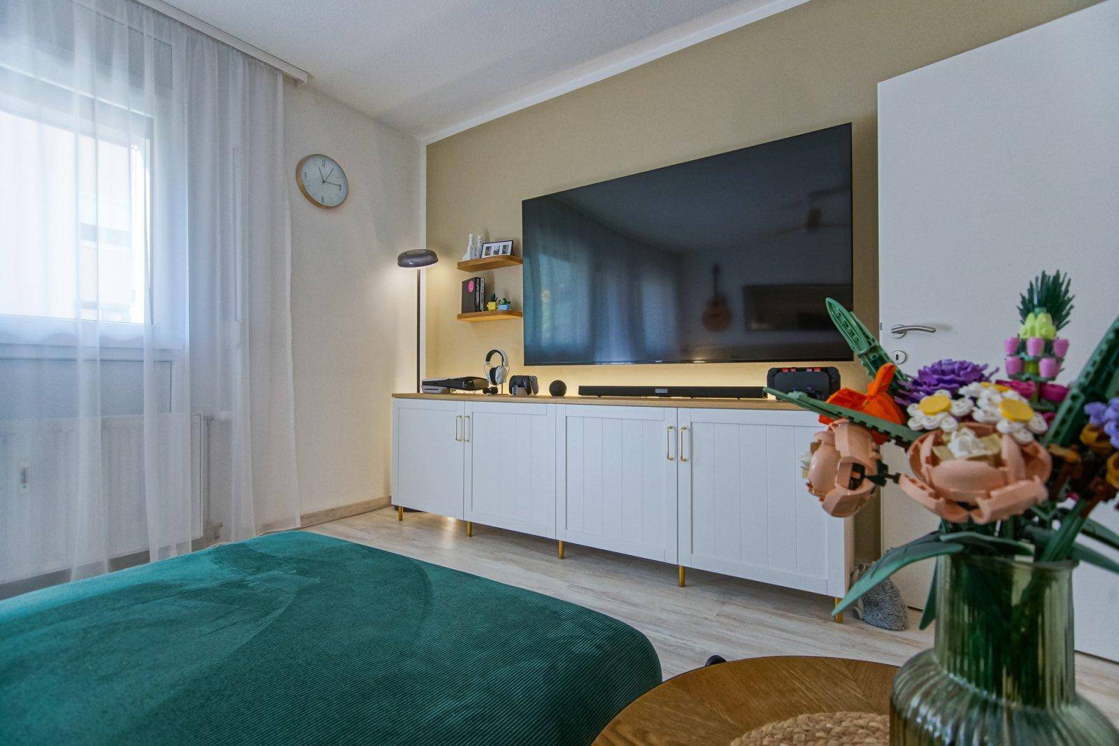 Wohnzimmer mit großem Fernseher, modernen weißen Schränken und hellem Fenster.