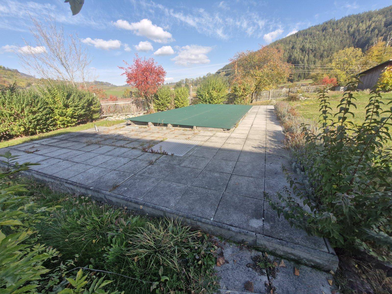 Gepflasterte Terrasse mit abgedecktem Pool, umgeben von grüner Bepflanzung.