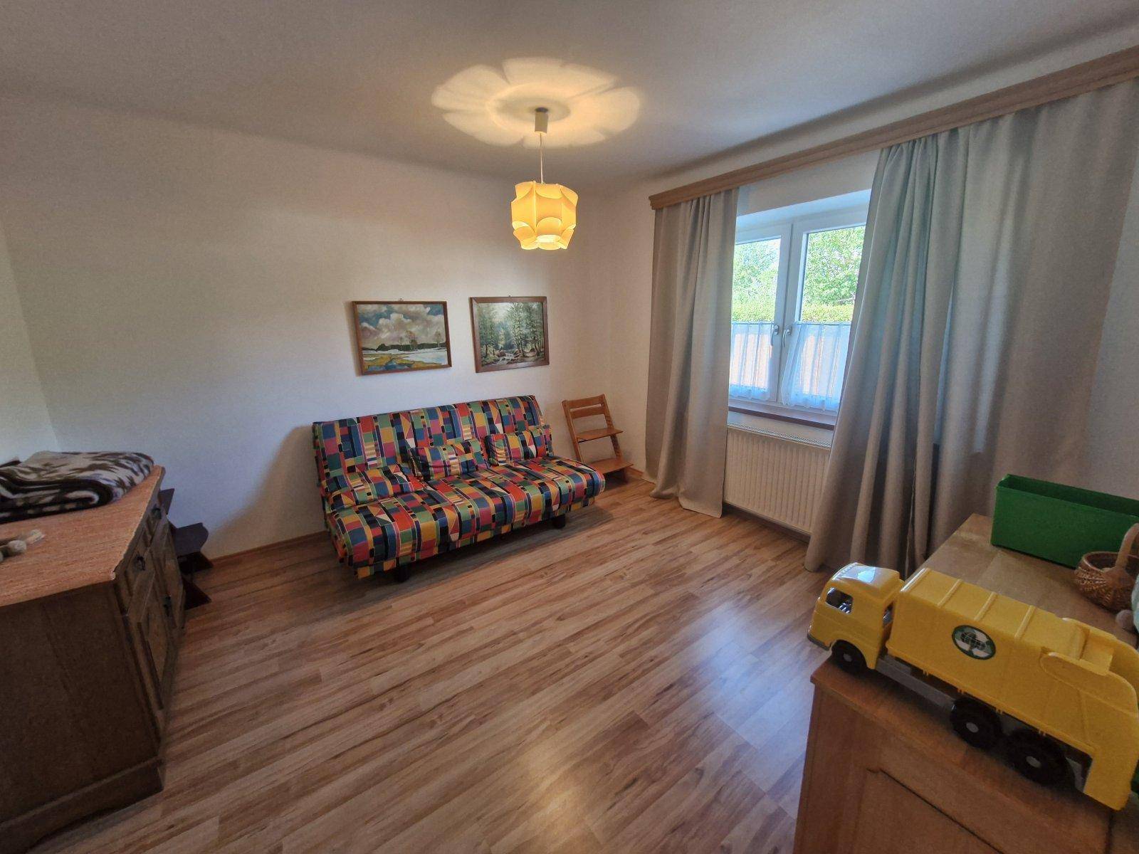 Helles Zimmer mit Holzboden, buntem Sofa und Fenster mit Blick ins Grüne.
