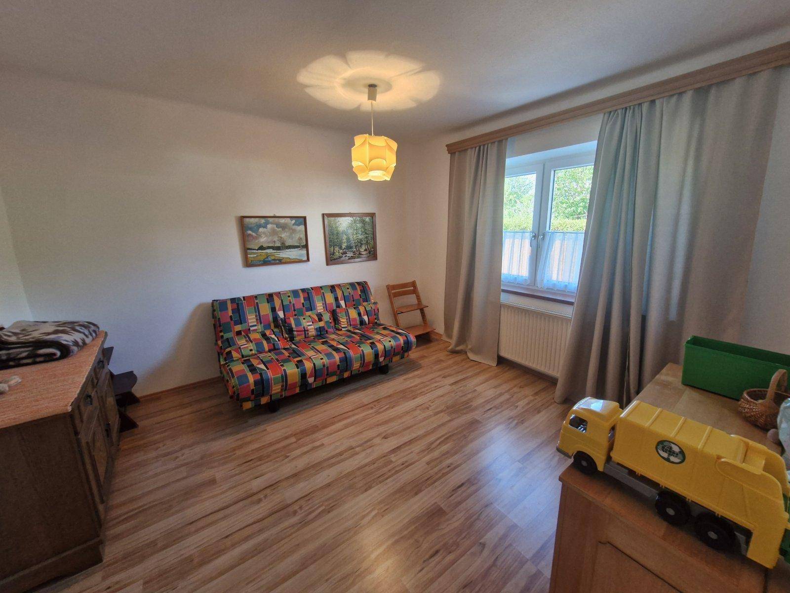 Helles Zimmer mit Holzboden, buntem Sofa und Fenster mit Blick ins Grüne.