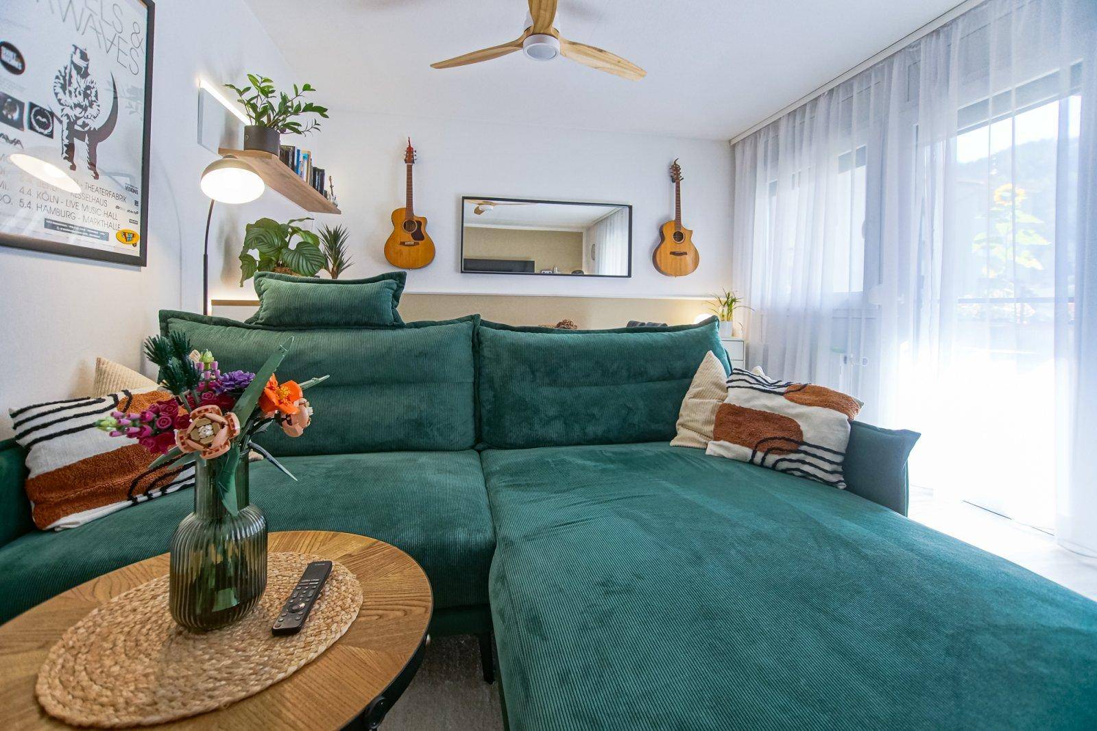 Helles Wohnzimmer mit grüner Eckcouch, Deckenventilator und großen Fenstern mit Bergblick.