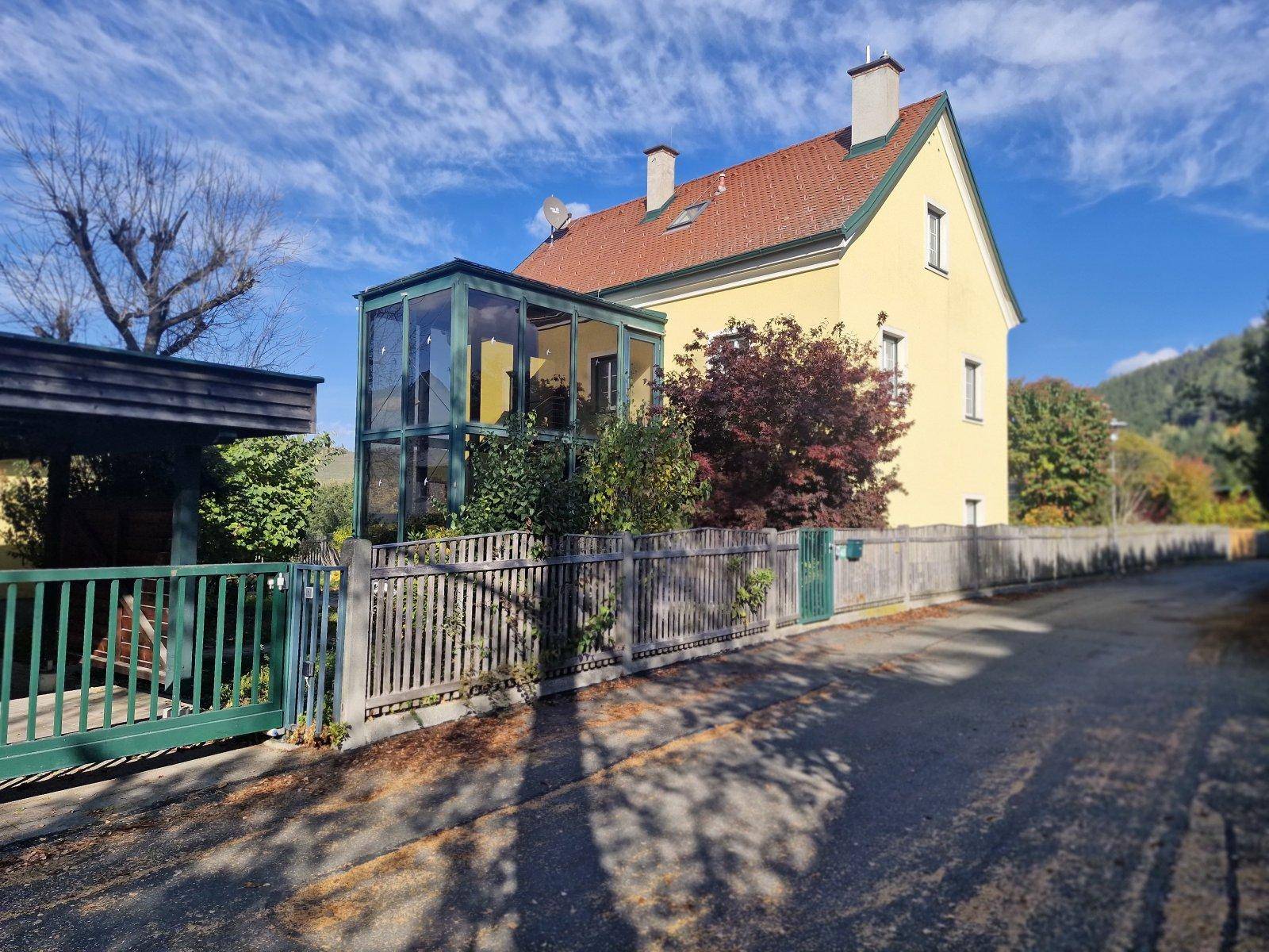 Ansicht des Hauses von der Straße mit Zaun, Carport und verglastem Anbau.