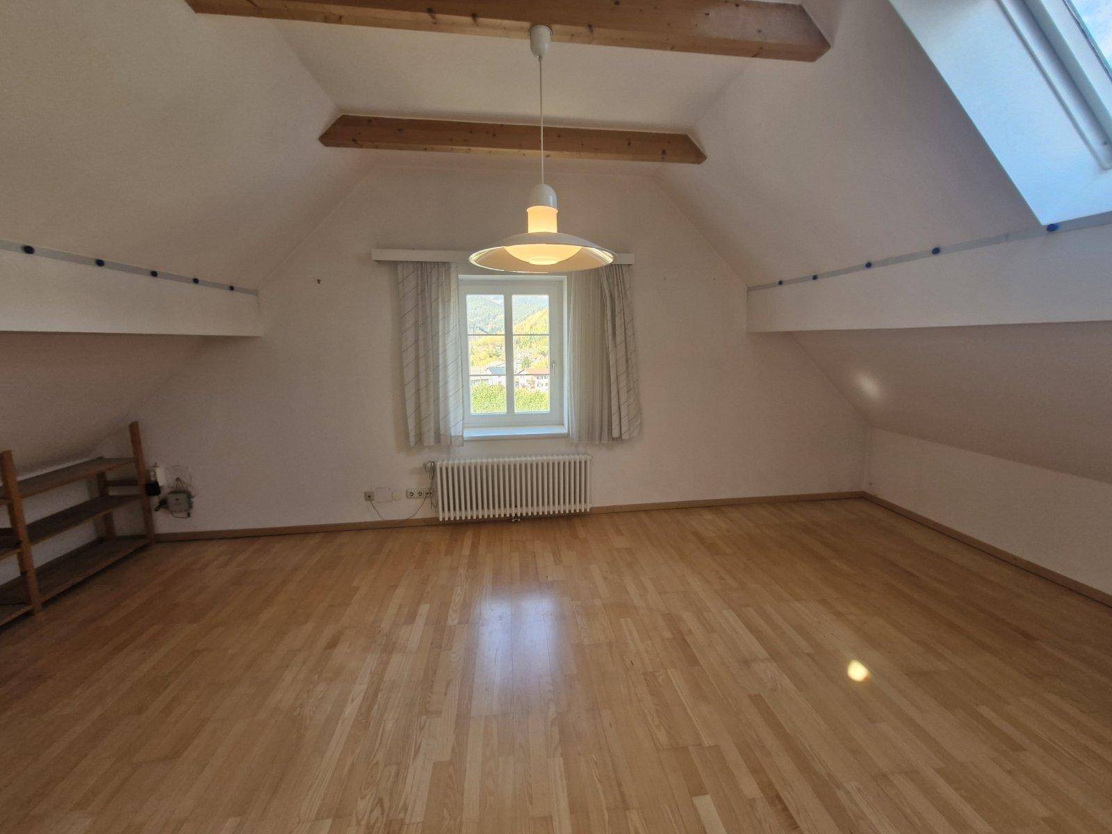 Dachgeschosszimmer mit Holzbalken, Parkettboden und einem Fenster mit Blick ins Grüne.