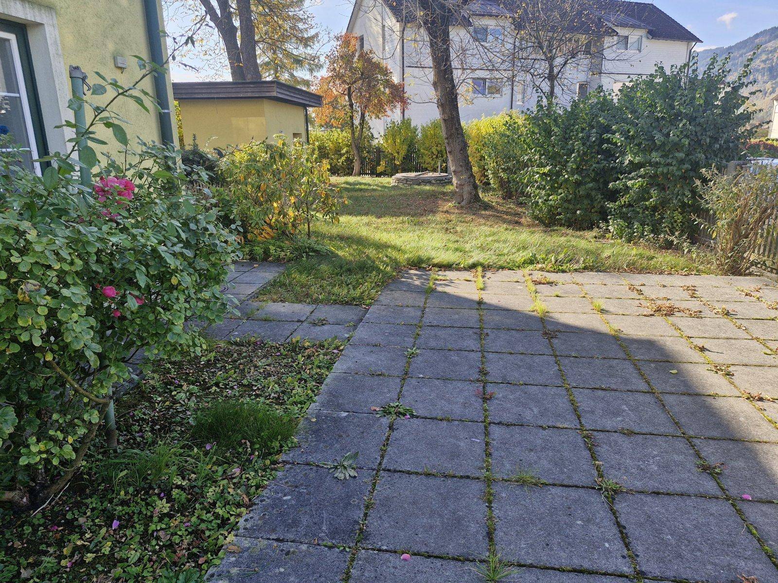 Gepflasterter Gartenweg umgeben von Blumenbeeten und Sträuchern in sonniger Lage.